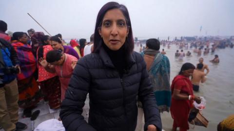 Samira Hussain wearing a black gilet standing with a crowd of people at the confluence of the Ganges and Yamuna rivers.