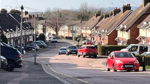 A residential street. There are cars parked outside of houses. Two of them are bright red. 