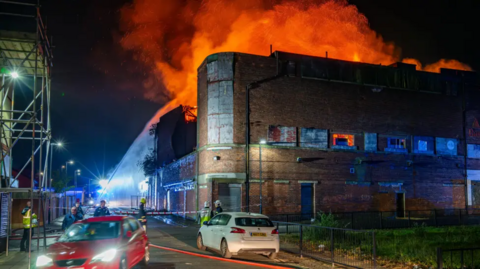 Massive orange blaze above a building and fire brigade spraying water at it. A red car and a white car are on the road next to it.
