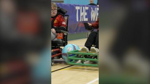 A blue football is wedged between two powerchairs being driven in a sports hall. A young boy is seen on the left wearing a Manchester United FC jersey.