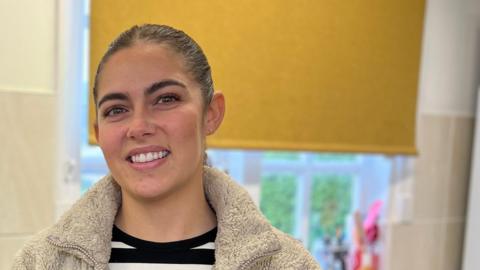 Molly smiles at the camera as she stands by a kitchen counter with two cake mixers to her left. She's wearing a white fleece with a black and white stripped t-shirt. Her hair is tied back in a ponytail.