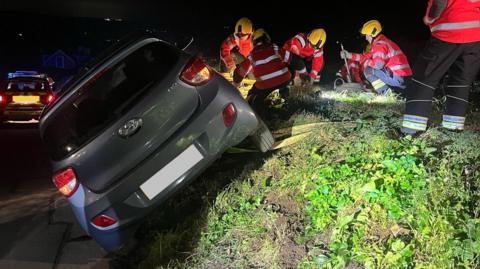 A grey car mounted on a mound of grass at a 45 degree angle, with fire officers standing on the mound holding onto the car by anchoring it with equipment.