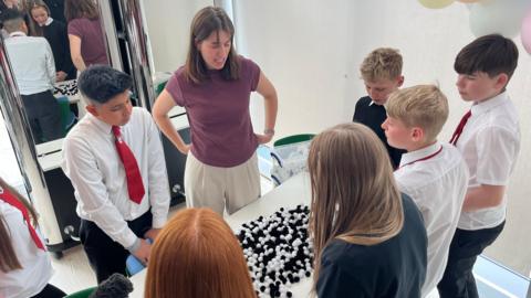 Seven young secondary school pupils and a teacher standing in front of desk which has some black and white balls on it which represent the HPV virus