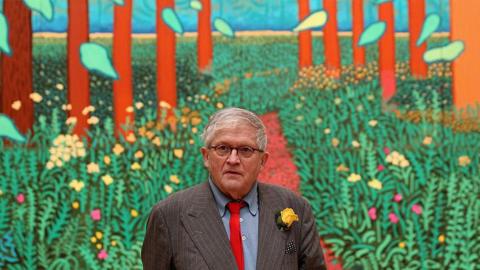 A man with grey hair and wearing black-rimmed glasses is standing in front of a colourful painting depicting green plants and tall trees. He is wearing a grey pin-striped suit, blue shirt and red tie with a yellow flower in his lapel. 