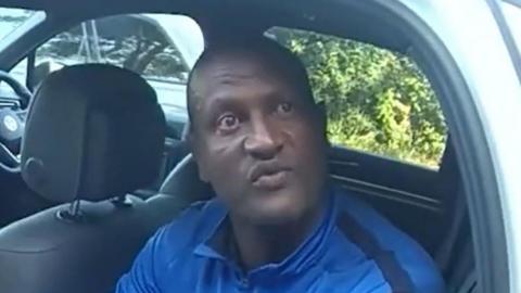 A still of Hadush Kebatu, taken from a video. He is wearing a blue quarter zip jumper while sitting in the back of a police car. He has his lips pouted while in the middle of talking.