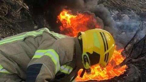 A firefighter in a fluorescent yellow and brown uniform is crouching in front of a bright orange flame. The firefighter is wearing a helmet as smoke billows from the scene.