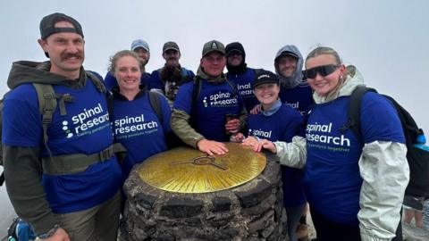 Sam Baldwin is stood atop Yr Wyddfa alongside family and friends. They are all touching the mountain peak, and they are wearing blue 'Spinal Research' t-shirts.