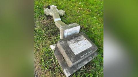 A gravestone shaped in a cross in a churchyard which has been smashed and fallen from the base on the grass.
