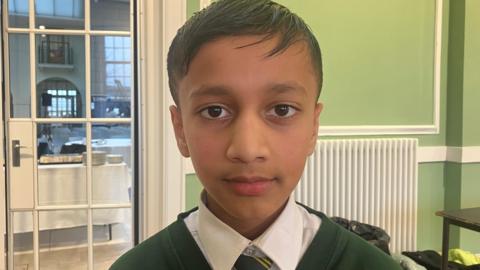 Soham, a pupil at St Malachy's Primary school in Belfast. He has short, dark hair and is wearing a dark green school jumper, white shirt and a green and yellow striped tie.