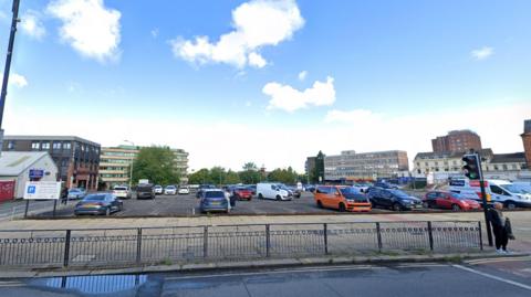 An open-air car park on a sunny day. There are high-rise buildings in the background, and a pedestrian is crossing the street in front of the car park.