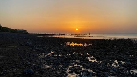The sun dips down in to the sea off the coast of Cromer. Groynes in the sea are in silhouette and there are rocks in the foreground. The sky has hues of orange and grey.