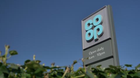 A co-op logo on a sign against a blue sky with a green hedge in the foreground