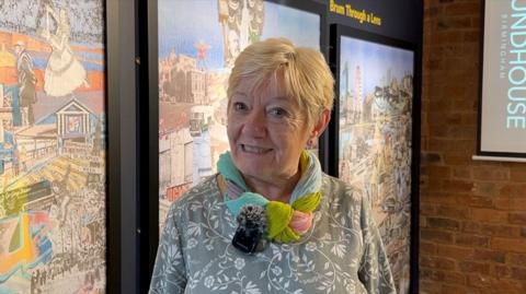 Betty Cowley who has short blonde hair and stands in front of designs showing the past around The Roundhouse. She wears a multi-coloured scarf above a grey top with a white floral design