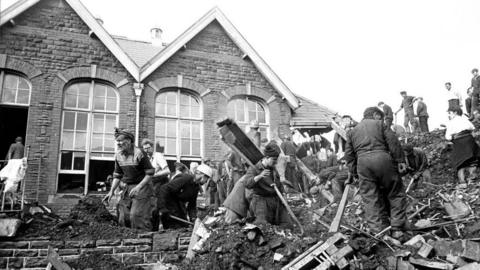 A black and white image of Pantglas school after the Aberfan disaster. Men are working through the rubble trying to clean up and rescue those trapped.