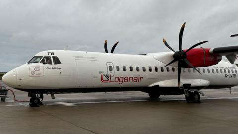 A white turboprop aircraft with a red tail and engine nacelles, parked on a wet airport apron under an overcast sky. The fuselage displays the word “Loganair” in large red letters with a stylized logo, and the text “Clan Donnachaidh” near the cockpit. The aircraft has two black propellers, and a ground power cable is connected to the nose gear area.