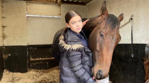 A woman with long dark hair tied back and wearing a coat, stands in a stable next to a brown horse.