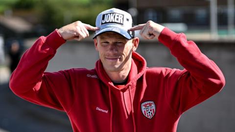 James McClean in a Derry City tracksuit pointing at his baseball cap