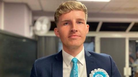 A head and shoulders shot of a man with short blond hair wearing a navy blazer, white shirt and light blue tie smiling into the camera. The background is blurred but he is stood inside a room with a fan in the corner.