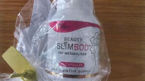 A clear plastic package laid on a wooden table with the a tub of the fake Slimbod pills inside. The packaging reads 'Beauty Slimbod Fat Metaboliser' - one months supply. It is white and pink in colour.