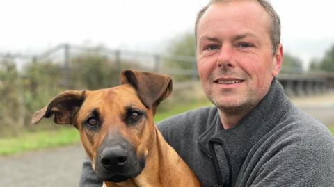 Ripley the dog, a tan coloured Dobermann cross, looks at the camera alongside owner Rob, who is smiling and wearing a grey jumper