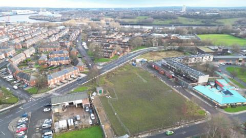 An aerial view of an area of Newcastle. In the foreground is a muddy field next to a row of flats. Much of the area is made up of terraced houses, many of which have solar panels on their roof. The River Tyne can be seen in the background.