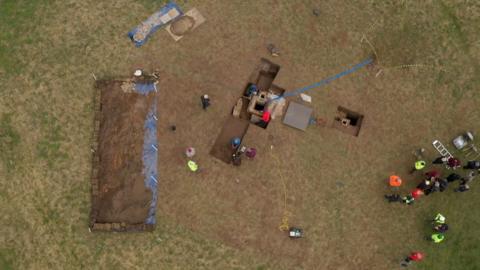 An aerial view of a field dug up with rectangular patterns. Groups of people in hi-vis stand around.
