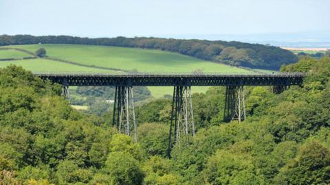 Meldon viaduct seen sideways on with trees around the base of the viaduct at both ends and fields stretching out across Dartmoor