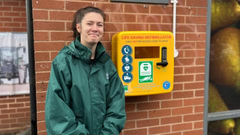 A woman with brown hair in a ponytail and dressed in a green paramedic jacket is smiling and stood against a yellow box fixed on a red brick wall. The yellow box says life saving defibrillator. Call 999 for access code to open.