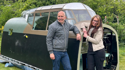 A picture of the cockpit, with Adrian and Belle smiling and leaning on it.