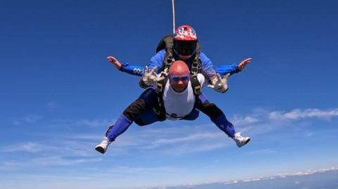 A man skydiving, with an instructor on his back. The man is bald and the instructor is wearing a helmet with a dark visor. There is blue sky around them.