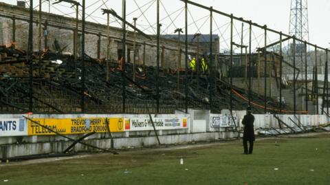 Charred remains of a football stand, strewn with tape. A single police officer looks on.