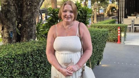 Julie Osgood, pictured in her late 50s, wearing a sleeveless white dress and with light blonde hair cut into a bob, stands in front of a green hedge and smiles at the camera.