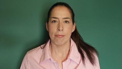A woman with brown hair looks at the camera. She is wearing a pink and white shirt, and is standing in front of a dark green wall. 