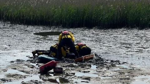 Mud with puddles on the surface with three RNLI crew members crouched together on the mud balanced on a stretcher to the right and a paddleboard to the left.