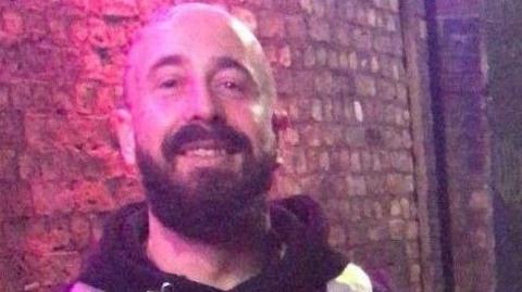 Donovan Travis Dineen-Whitham smiles at the camera. He is bald with a brown beard. He is standing in front of a wall.