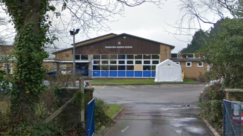 Google Street View of Ferndown Middle School viewed through the gate from the road. The single storey building has a pitched roof and blue cladding beneath the windows.