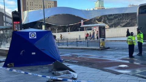blue police tent on a pavement, cordoned off by blue and white police tape. Two police officers are standing inside the tape wearing black hats and yellow hi-vis jackets. a bus drives past