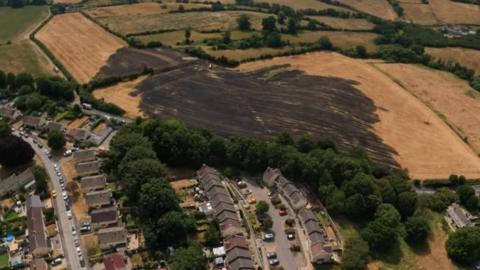 A blackened, scorched field in the countryside surrounded by brown and pale green fields behind and to the side. In front of the burnt field is a road lined by green trees and in front of that are residential roads and houses.