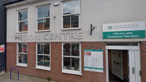 A two-storey building with the ground floor in brick and grey render on the upper floor. There are six windows and a door, which has a sign reading: Library and Customer Services above it.