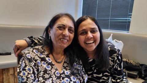 Prevanda Varshani (left) and Dishna Hirani (right) sit together hugging and smiling for a photo