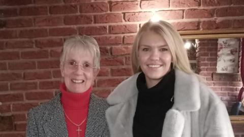 Sue, wearing a red turtle-neck jumper and a blac and white coat, next to Chloe Ball, wearing a black jumper with a grey fur coat