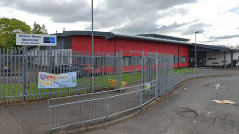 A school, with buildings coloured red and white. Gates lead into both the school and a car park nearby. 