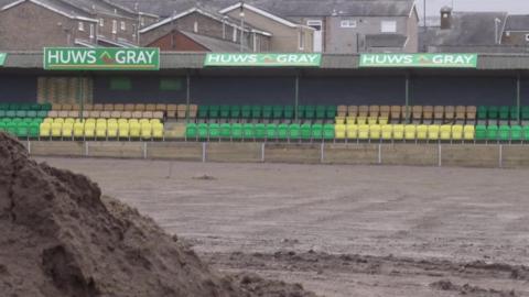 Eisteddle presennol yn safle CPD Caernarfon gyda stad dai yn y cefndir. Yn y blaen mae rhan o gae'r Oval sydd yng nghanol cael ei lefelu fel rhan o gynllun uwchraddio ac mae tomen o bridd yng nghornel chwith gwaelod y llun.