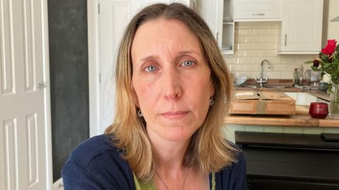 Lisa Lailey looking at the camera in front of her kitchen. Lisa has blonde hair and blue eyes. She is wearing a green top with a navy cardigan.