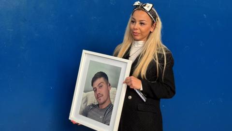 Eden Hollis, a woman with long blonde hair a black and white bow in her hair, a white top, black coat and she is holding a white framed photo of Lloyd Donkin who has short brown hair, stubble, grey top and is sitting on white sofa.