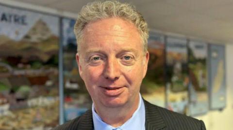 Alex Allinson, who has short, fair, wavy hair, is wearing a dark pinstriped suit jacket and light blue shirt.