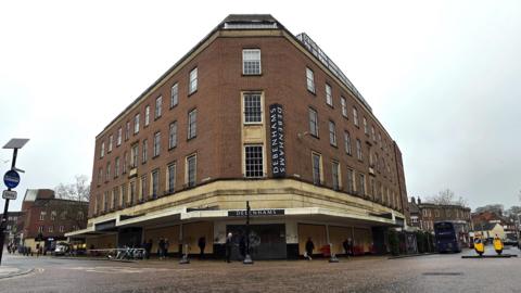 The exterior of the Debenhams building in Norwich on a grey day. It is a multi-storey building, constructed in the 1950s. Shoppers are walking past it.