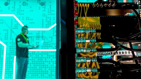 A man standing against a bright green background in a data centre - in the foreground is the back of a large computer with lots of yellow and black wires.