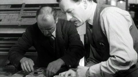 Black and white image of Mr Thomas in the foreground in a shirt, tie and wastecoast, piecing together the layout of the newspaper with another man