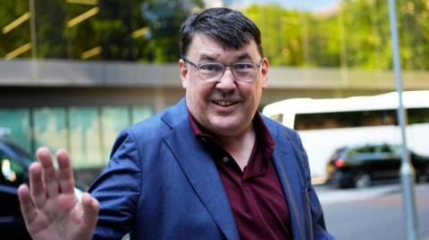 Linehan smiles and waves at the camera while standing outside in front of a road 